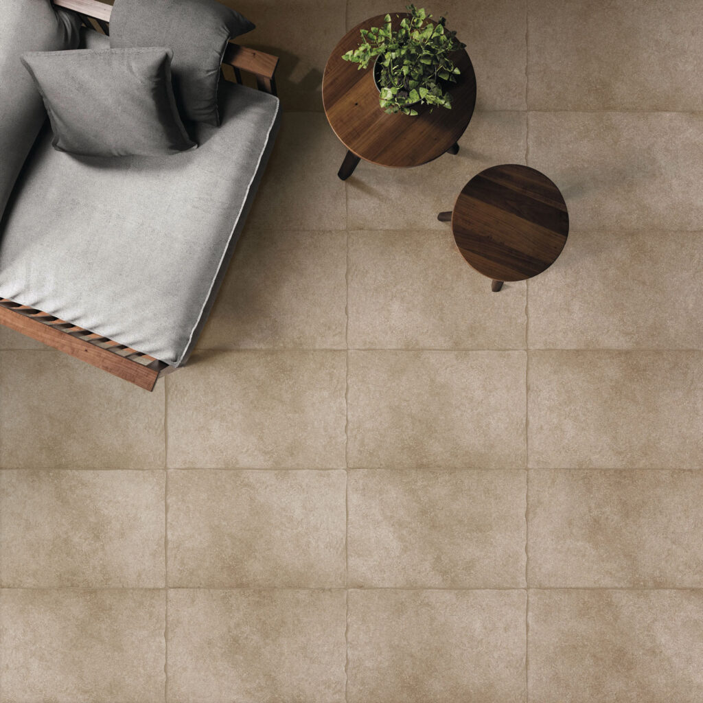 Vista dall'alto di un ambiente outdoor con pavimento in gres porcellanato effetto pietra beige a grandi lastre squadrate. Sono presenti un divano moderno con cuscini grigi e due tavolini circolari in legno scuro con una pianta ornamentale.