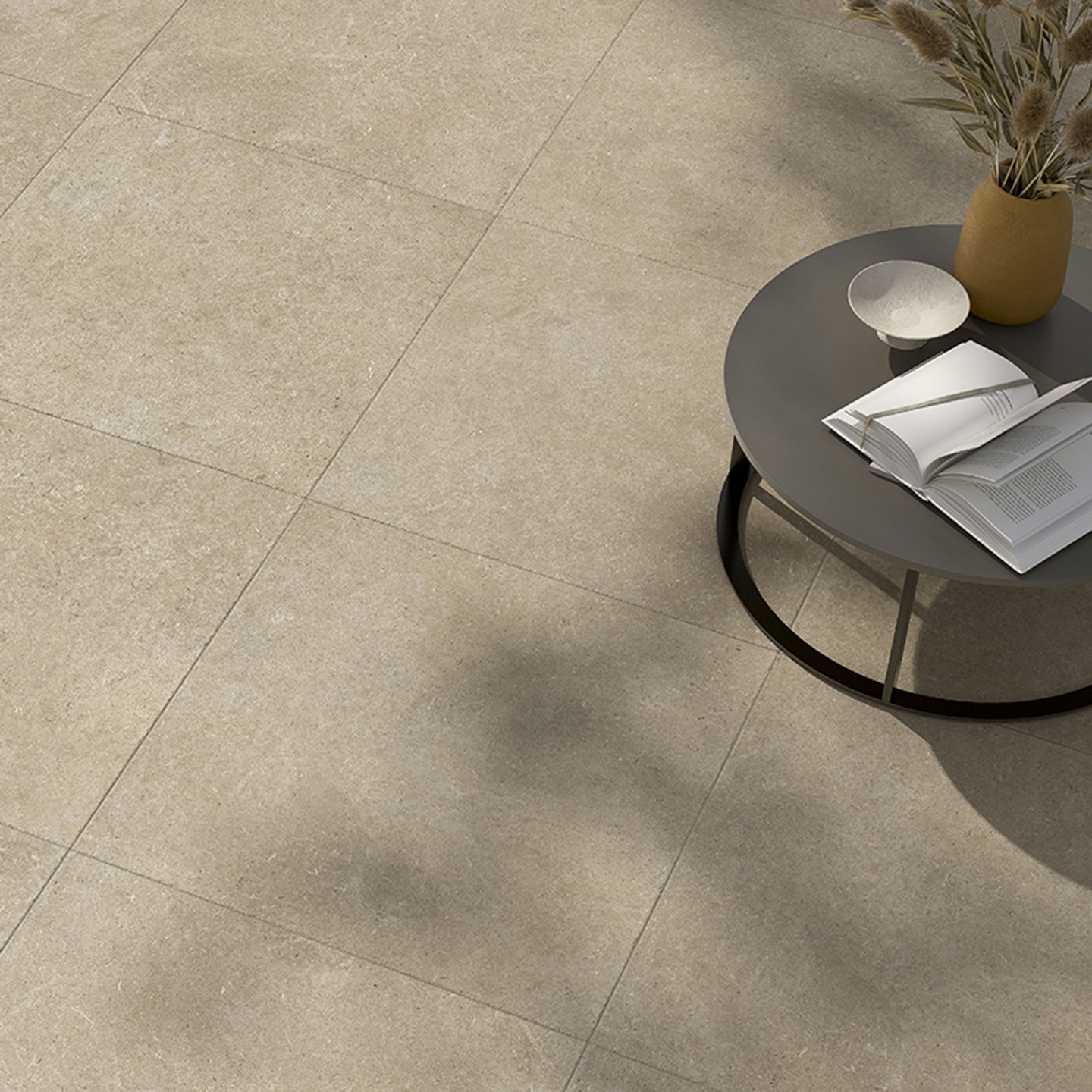 Dettaglio di una pavimentazione interna in piastrelle di gres porcellanato color crema con texture naturale. L'ambientazione minimalista include un tavolino basso in metallo scuro con libri e un vaso di fiori secchi, illuminata da una luce solare calda.