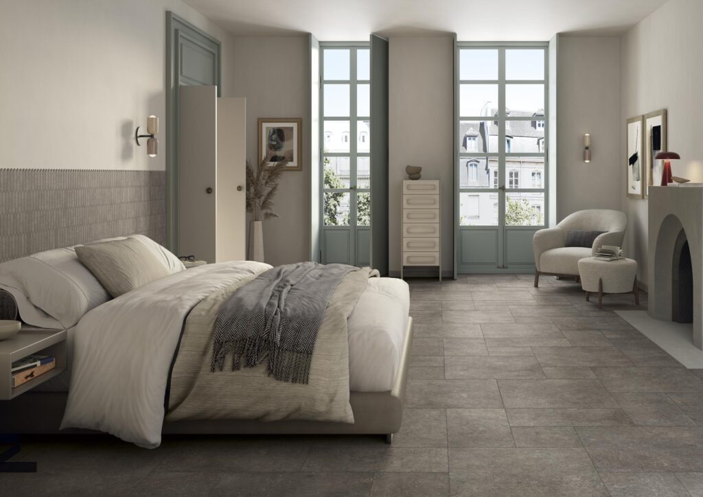 Collezione Cathédrale, variante Amiens Acier. L'ambiente è una camera da letto moderna dove il pavimento grigio scuro si sposa con una parete decorata a mosaico allungato e arredi minimalisti.
