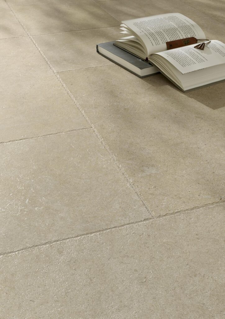 Dettaglio della collezione Cathédrale, colore Paris Crème. Piastrelle in gres porcellanato color crema con texture naturale, sulle quali sono appoggiati libri aperti in un contesto di design raffinato.