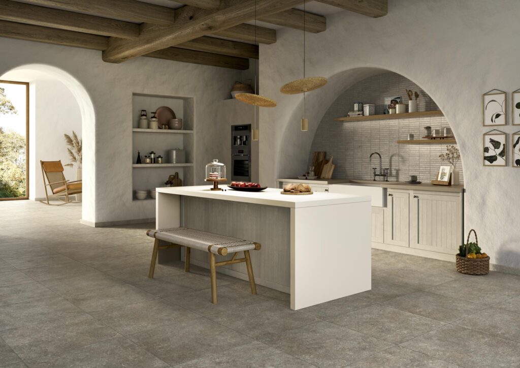 Cucina rustica di design con pavimento della collezione Cathédrale, variante Rouen Corde nel formato 60x60. L'ambiente presenta travi in legno a vista, un'isola centrale bianca e piastrelle grigie calde.
