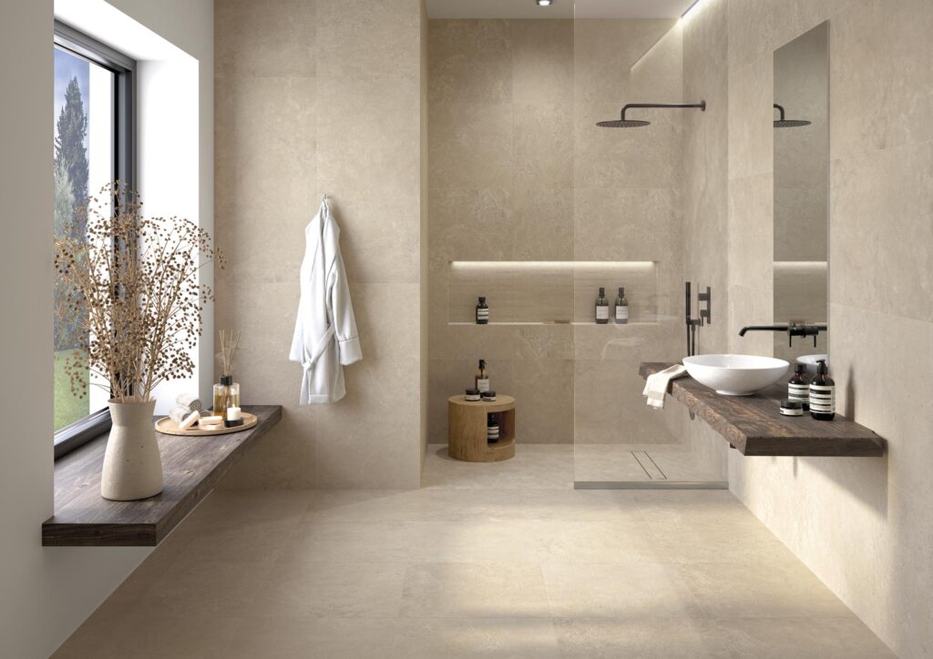 Collezione G Stone, variante Dordogne Lin: un raffinato ambiente bagno caratterizzato da piastrelle in gres porcellanato effetto pietra in grandi formati. La scena mostra una posa armoniosa e continua tra pavimento e pareti nei toni caldi del beige, completata da una moderna doccia walk-in con rubinetteria nera e un elegante piano lavabo in legno scuro, che insieme creano un’atmosfera spa luminosa e naturale.