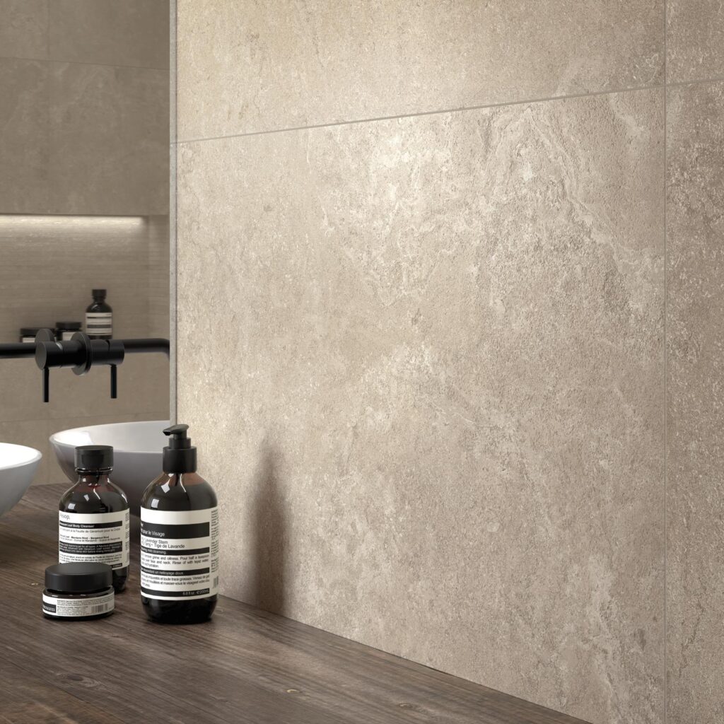 Collezione G_Stone nella variante cromatica Dordogne Lin: un raffinato rivestimento per l'ambiente bagno che riproduce la texture materica della pietra naturale in una calda tonalità beige. L'immagine mette in risalto la posa a parete a grandi formati, caratterizzata da venature delicate e una superficie tattile, abbinata armoniosamente a un piano lavabo in legno scuro, rubinetteria nera moderna e accessori di design per un'atmosfera spa contemporanea.