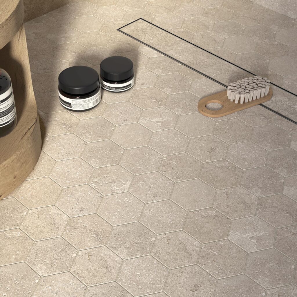La collezione G Stone arreda un bagno raffinato con un mosaico esagonale effetto pietra nella calda variante cromatica Dordogne Lin. L'immagine mostra il dettaglio di un pavimento doccia moderno con scarico a filo, dove le trame materiche delle piastrelle beige si armonizzano con accessori in legno e flaconi scuri, creando un'atmosfera di benessere naturale e design minimalista.