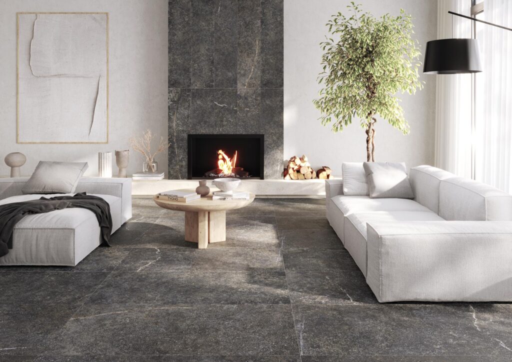 Collezione G-Stone nella variante Nero Ragusa: un raffinato ambiente living che mette in risalto l'eleganza del gres porcellanato effetto pietra scura. La pavimentazione a grandi formati e il rivestimento verticale del camino creano un dialogo armonioso con i luminosi divani bianchi e gli arredi minimalisti in legno, definendo uno spazio contemporaneo e accogliente impreziosito da un focolare acceso e luce naturale diffusa.