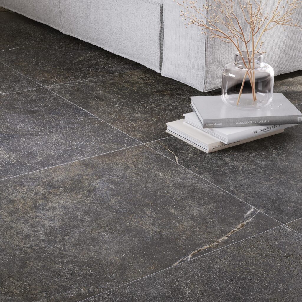 Collezione G Stone, pavimentazione in gres porcellanato effetto pietra nella variante Nero Ragusa, ideale per un ambiente living raffinato e contemporaneo. L’immagine mette in risalto piastrelle in grande formato con texture materica e venature naturali, posate con precisione accanto a un divano moderno. L'atmosfera è completata da elementi di design come libri d'arte e un vaso in vetro minimalista, che esaltano l'eleganza cromatica del nero antracite.