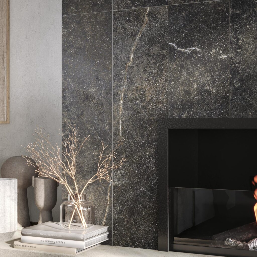 Collezione G Stone in variante Nero Ragusa: un raffinato rivestimento in grandi lastre effetto pietra dalle venature naturali, posato verticalmente in un ambiente living contemporaneo. L'immagine evidenzia la matericità della superficie ceramica accanto a un camino moderno, valorizzata da elementi d'arredo minimalisti come vasi di design, libri d'arte e rami decorativi che creano un'atmosfera lussuosa e accogliente.